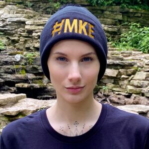 Woman wearing a black beanie hat with big old gold lettering of # M K E.