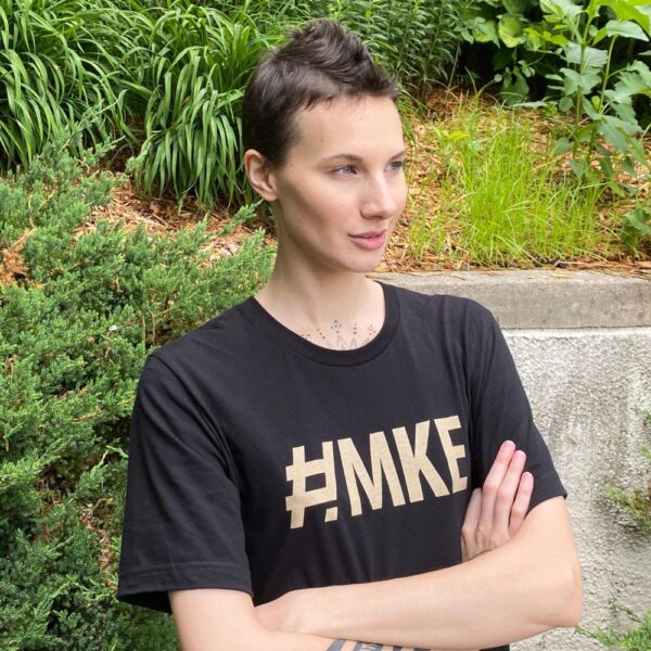 Woman posing with her arms crossed while wearing a black shirt with # M K E on the front in an old gold color.