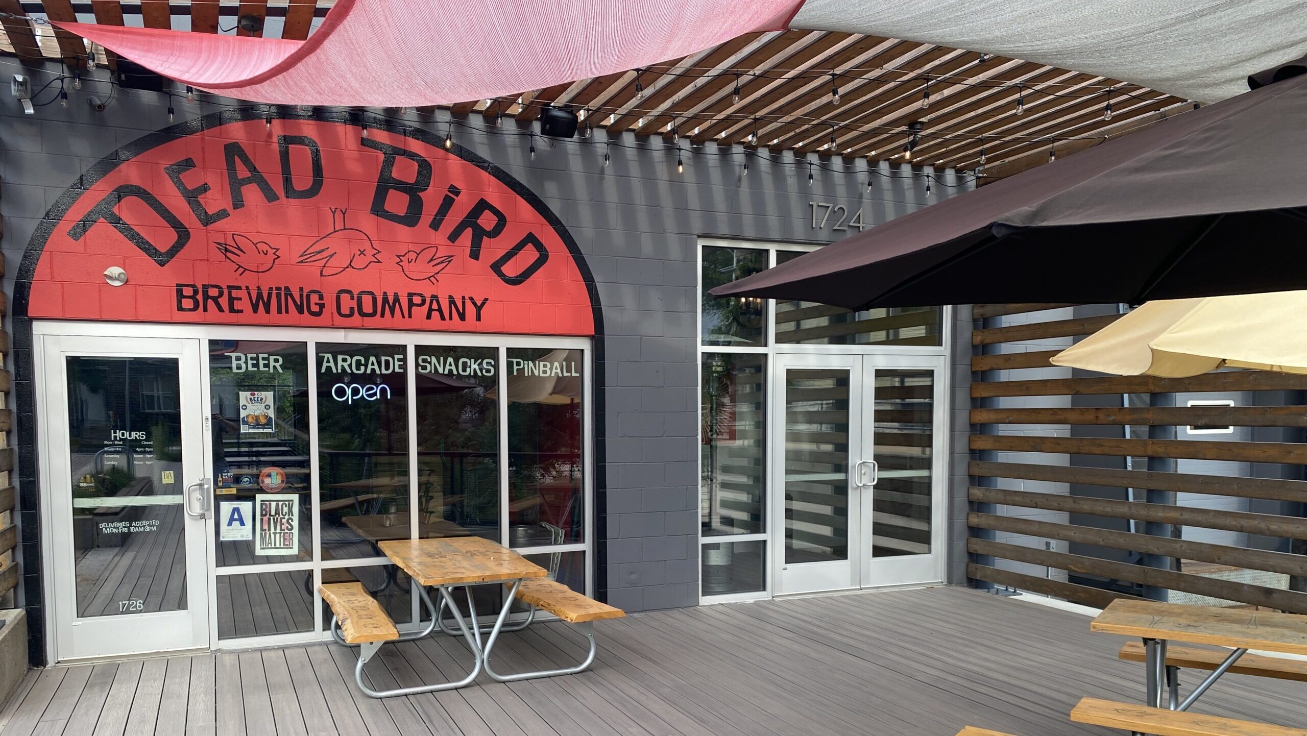 Exterior view of Dead Bird Brewing Company's entrance with a red canopy providing shade for picnic tables below.